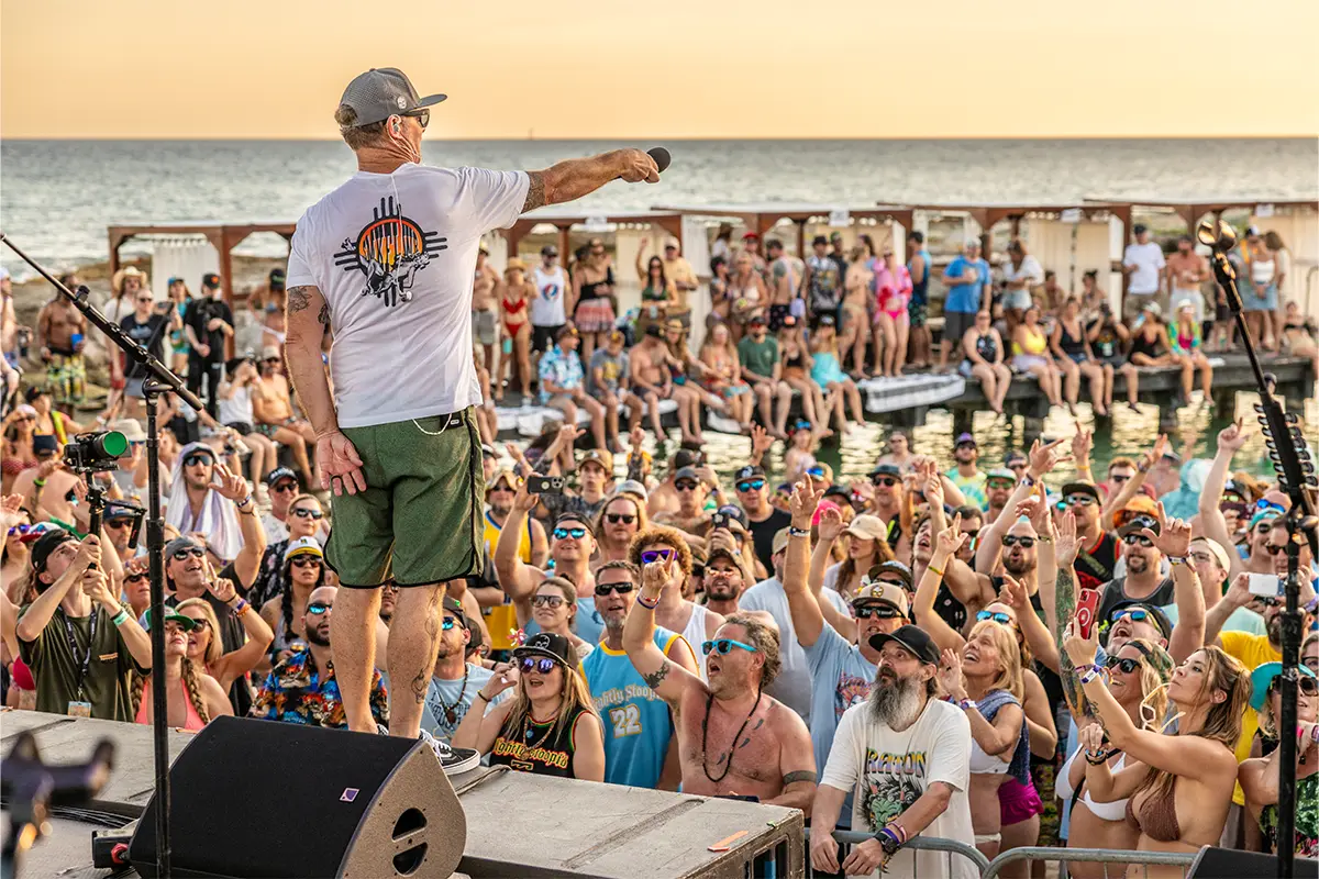 Miles from Slightly Stoopid holding his microphone to the crowd on the Beach Stage. Golden Hour sunset and ocean view in the background.